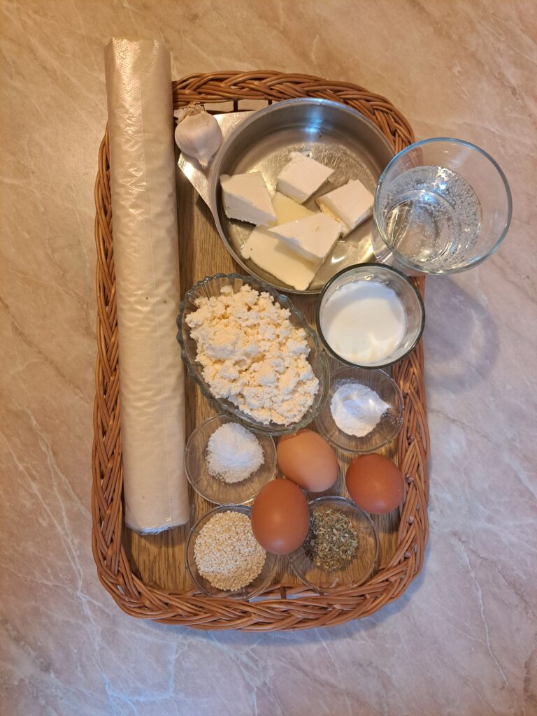 All ingredients for Cheese Phyllo Pie Bake arranged on a table, including phyllo sheets, cheese, eggs, yogurt, butter, oil, garlic, oregano, sesame, and baking powder.