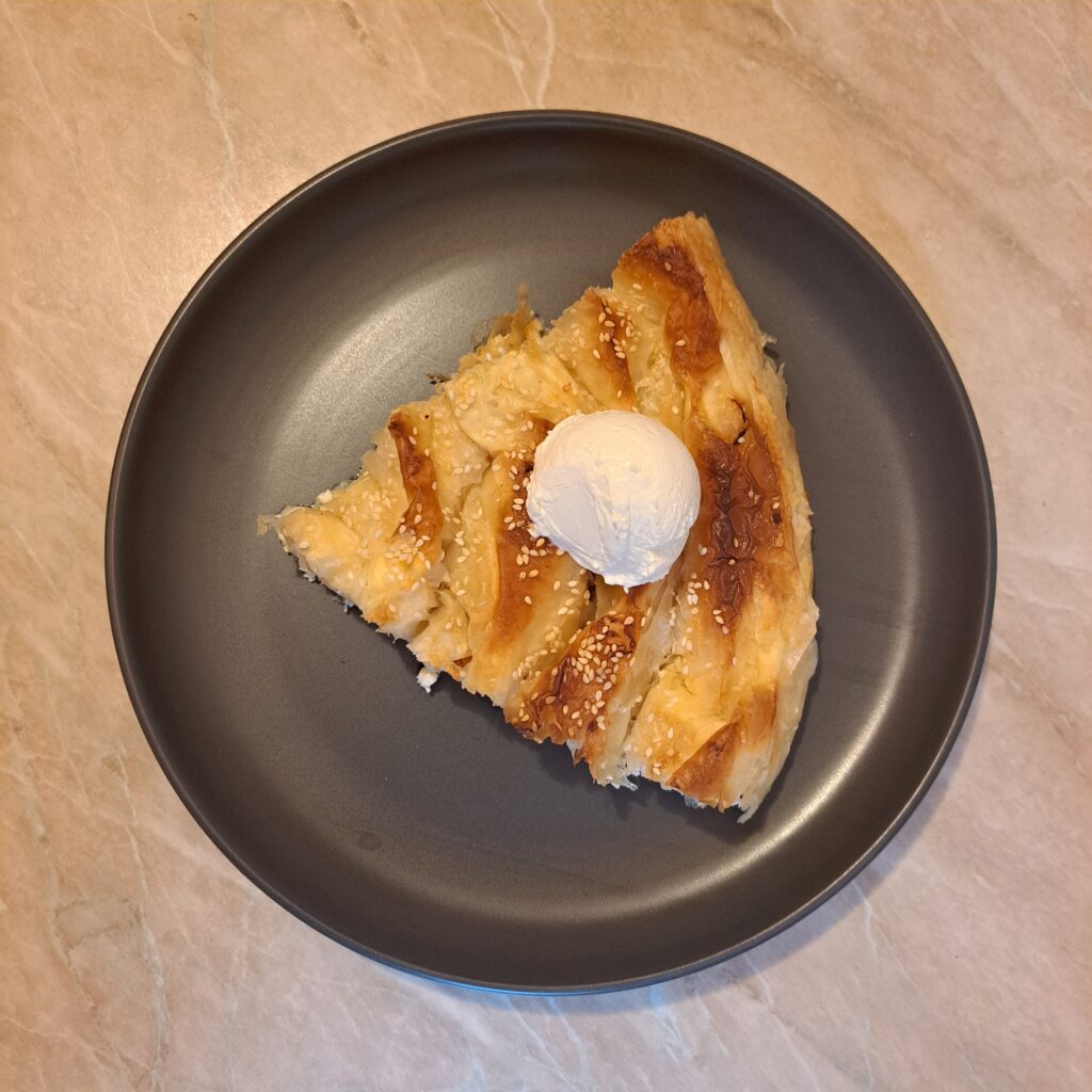 A served slice of Cheese Phyllo Pie Bake on a black plate, topped with a ball of sour cream and sprinkled with sesame.