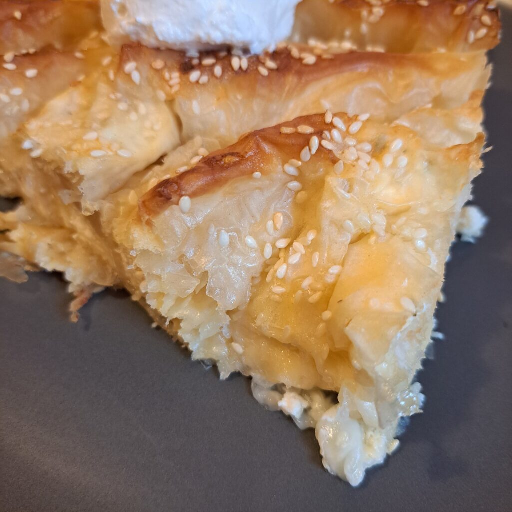 Close-up of a single slice of Cheese Phyllo Pie Bake on a black plate, topped with a ball of sour cream and sprinkled with sesame.
