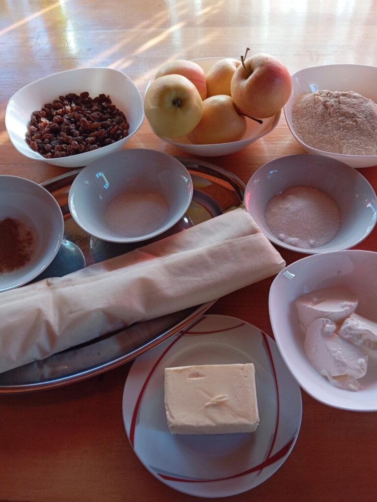 Ingredients for apple raisin phyllo strudel arranged on a table, including apples, raisins, phyllo sheets, butter, sugar, cinnamon, breadcrumbs, and sour cream.