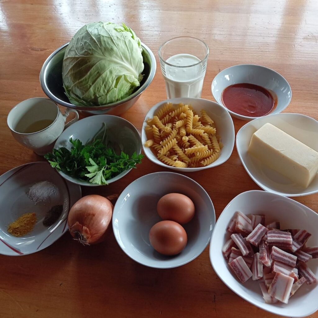 All ingredients for Creamy Cabbage Pasta Bake arranged on a table, including cabbage, pasta, pancetta, cheese, cream, eggs, and seasonings.