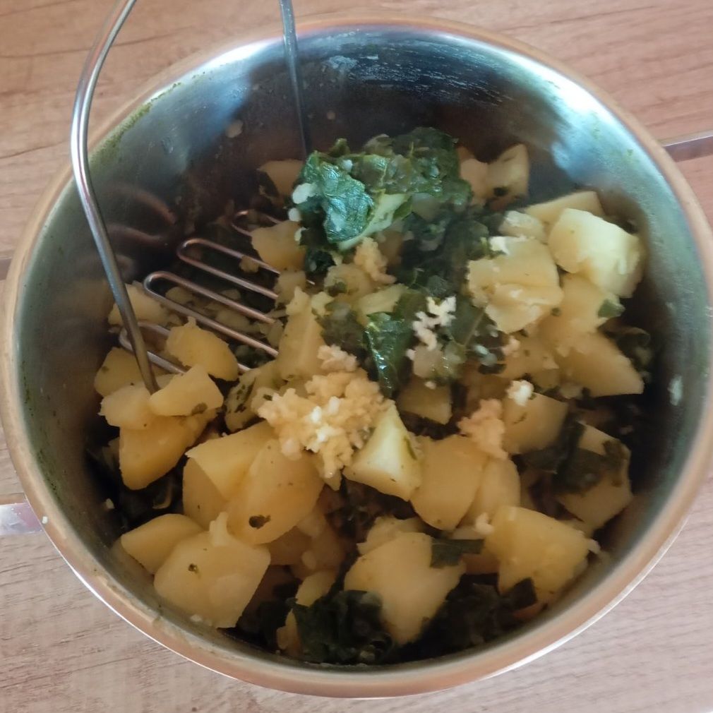 Close-up of mashed potatoes and collard greens being mixed smoothly in a bowl with crushed garlic and olive oil.