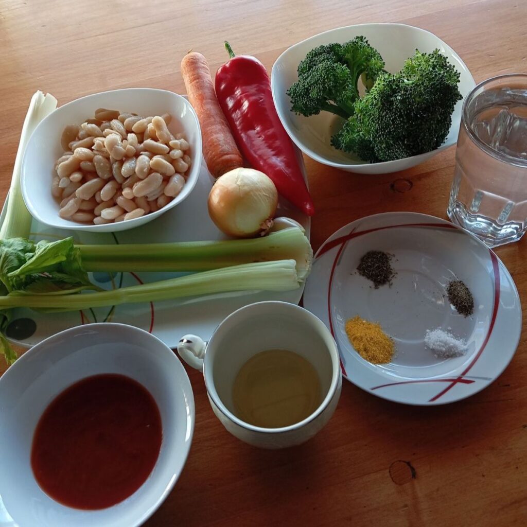 All ingredients for Hearty Vegetable Bean Soup arranged on a table, including onion, leek, carrot, celery, red pepper, broccoli, beans, garlic, tomato paste, oil, and seasonings.