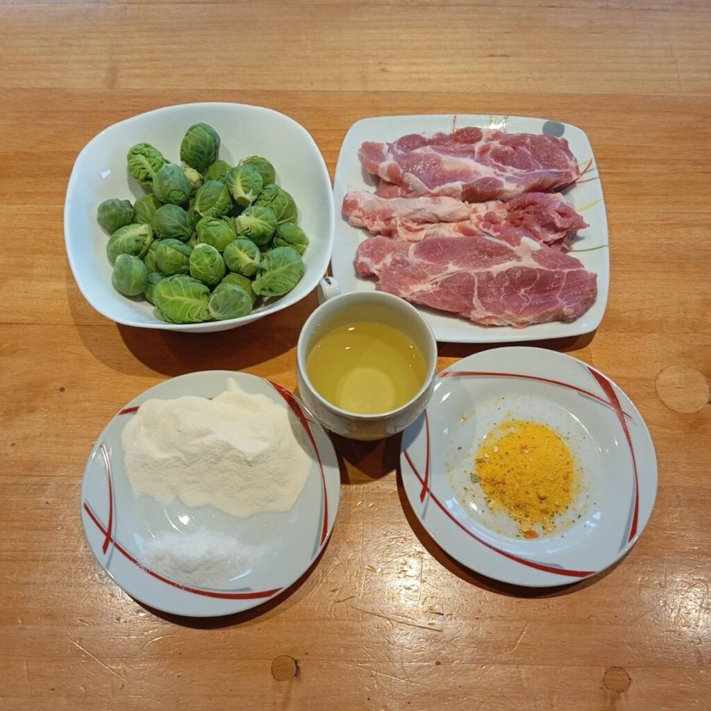 All ingredients for Brussels Sprouts Pork Schnitzels arranged on a table, including pork schnitzels, Brussels sprouts, flour, salt, seasoning, and cooking oil.