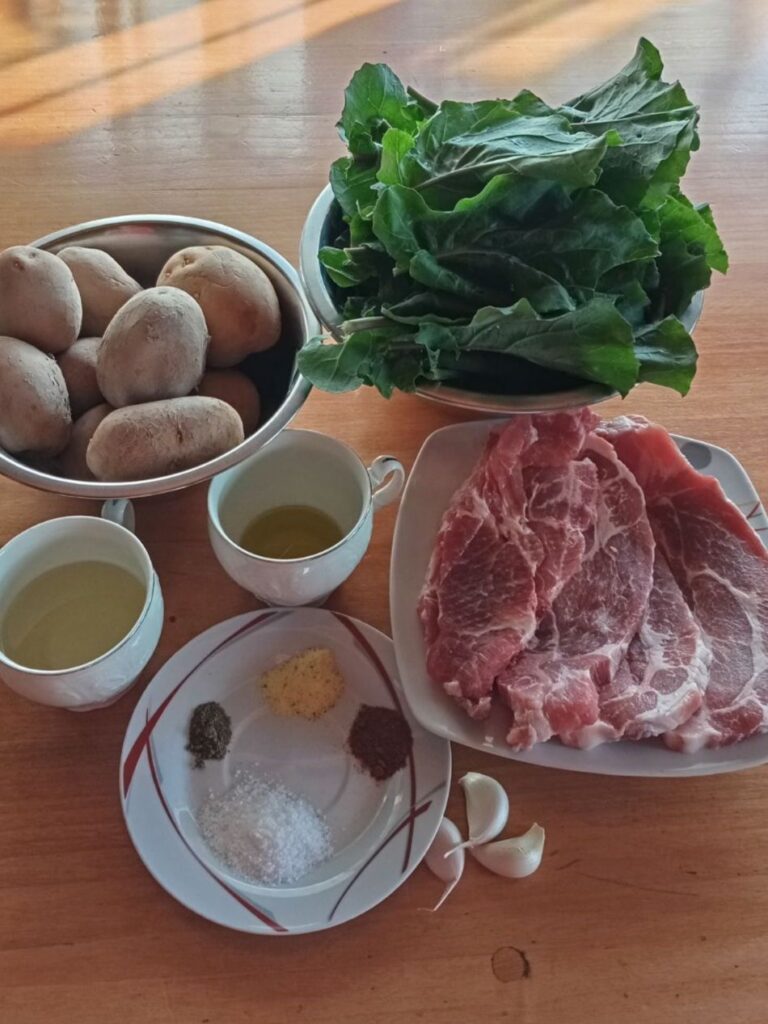 Ingredients arranged on a table including potatoes, collard greens, boneless pork neck chops, garlic, oils, and seasonings.