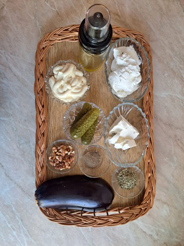 Ingredients for Creamy Eggplant Walnut Salad arranged on flat woven wooden basket, including eggplant, pickles, feta, sour cream, mayonnaise, walnuts, oregano, and black pepper.