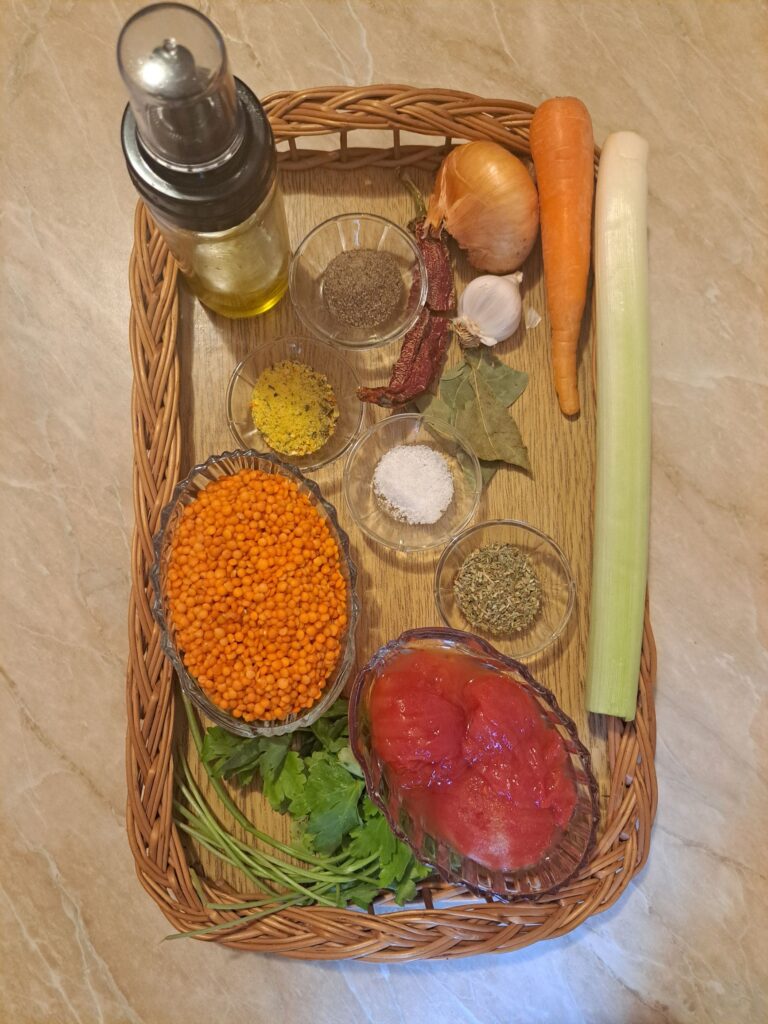 Top view of all ingredients arranged on a surface, including red lentils, canned tomatoes, leek, onion, carrot, garlic, spices, and cooking oil.