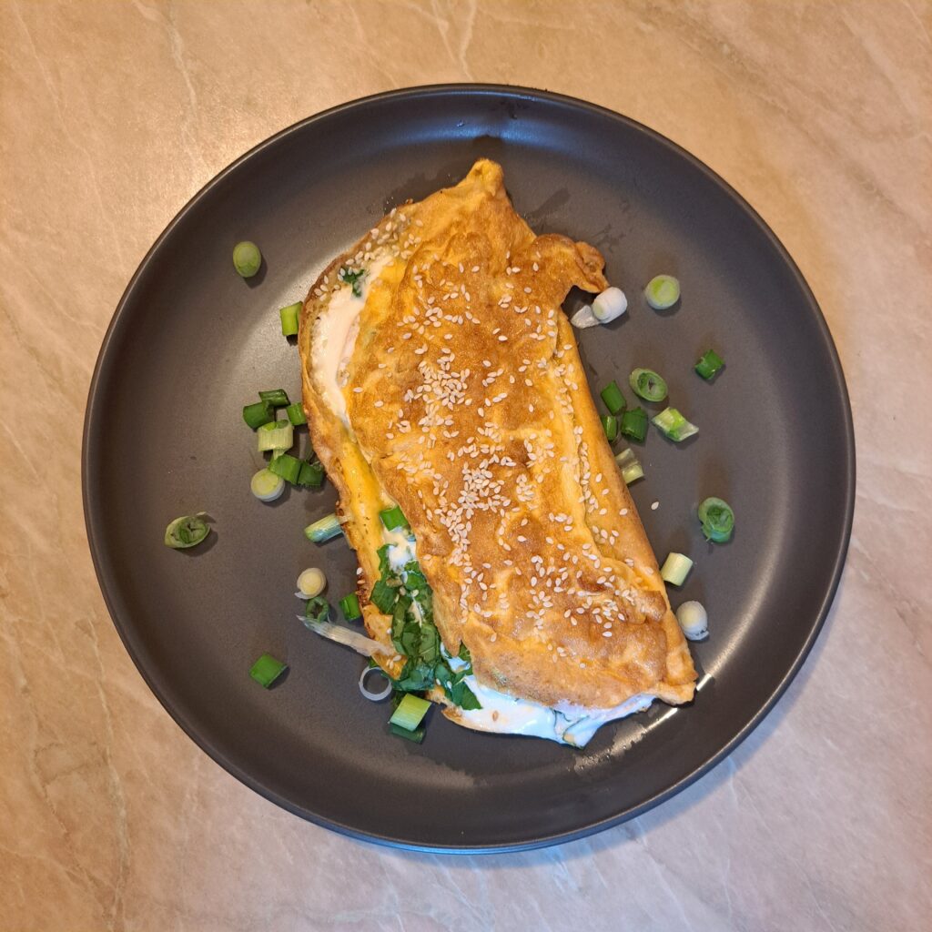 Freshly served Creamy Mushroom Arugula Omelette placed on a flat black plate, folded neatly, lightly sprinkled with sesame, and garnished with finely chopped young scallion.