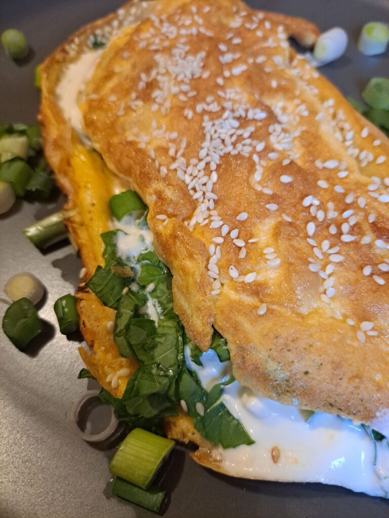 Freshly cooked creamy mushroom arugula omelette served on a flat black plate, lightly sprinkled with sesame and topped with chopped scallions.