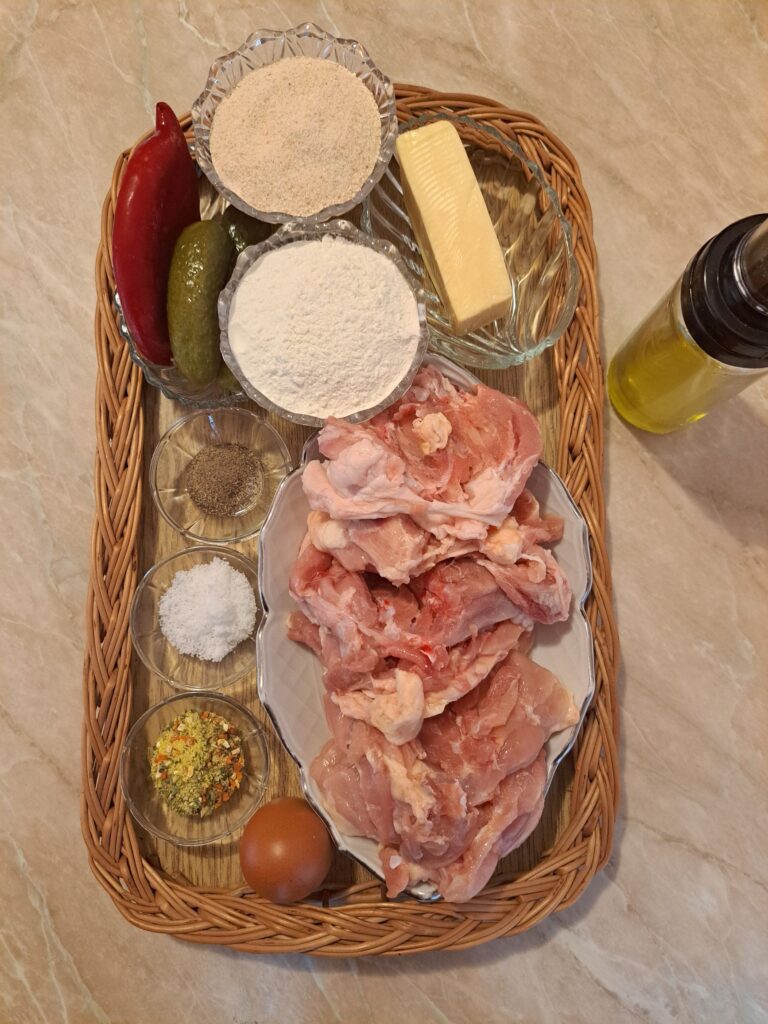 All ingredients for Crispy Stuffed Chicken Thighs arranged neatly on a flat woven wooden tray, including boneless chicken thighs, cheese, pickles, red pepper, egg, flour, breadcrumbs, and spices.