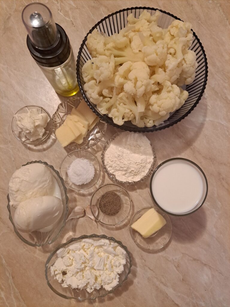 All ingredients for Creamy Cheesy Cauliflower Bake displayed on a table, including cauliflower, cheeses, milk, butter, flour, garlic, salt, and black pepper.