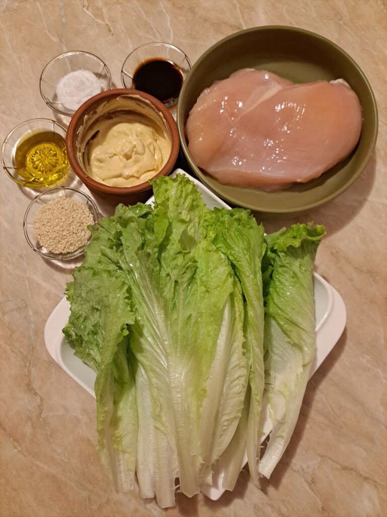 All ingredients for Grilled Chicken Lettuce Salad arranged on a table, including chicken breast, fresh lettuce, sour cream, mustard, sesame seeds, balsamic vinegar, olive oil, and salt.