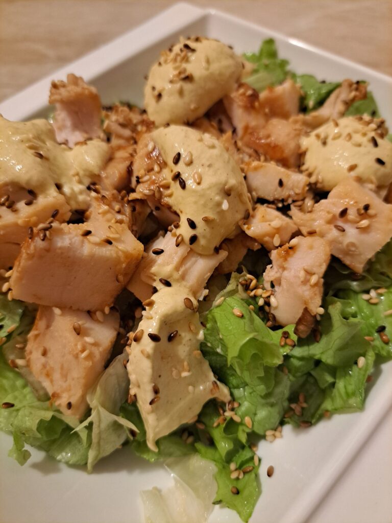 Close-up view of grilled chicken slices served on fresh chopped lettuce with creamy mustard dressing and toasted sesame on top.