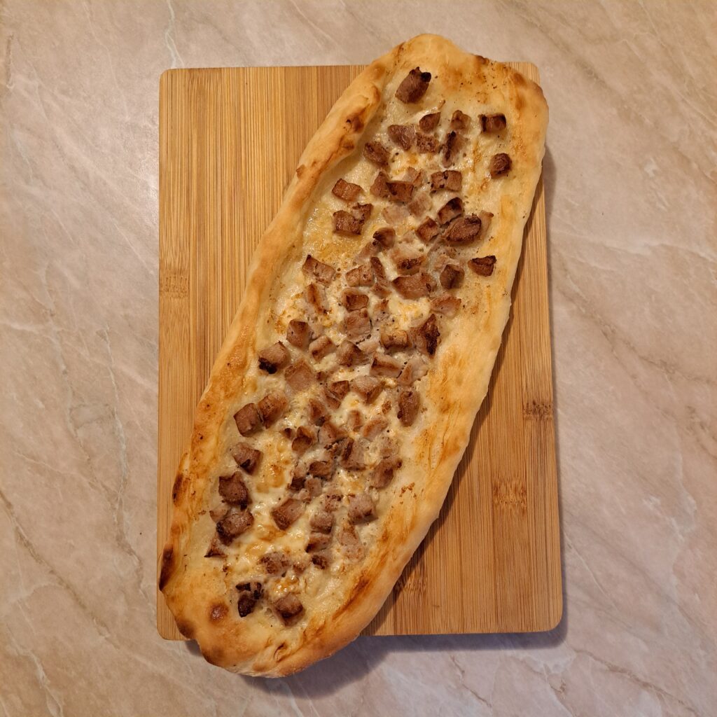 Whole Homemade Cheesy Pork Pastrmajlija served on a wooden board, showing golden crust, melted cheese, and evenly distributed pork cubes.