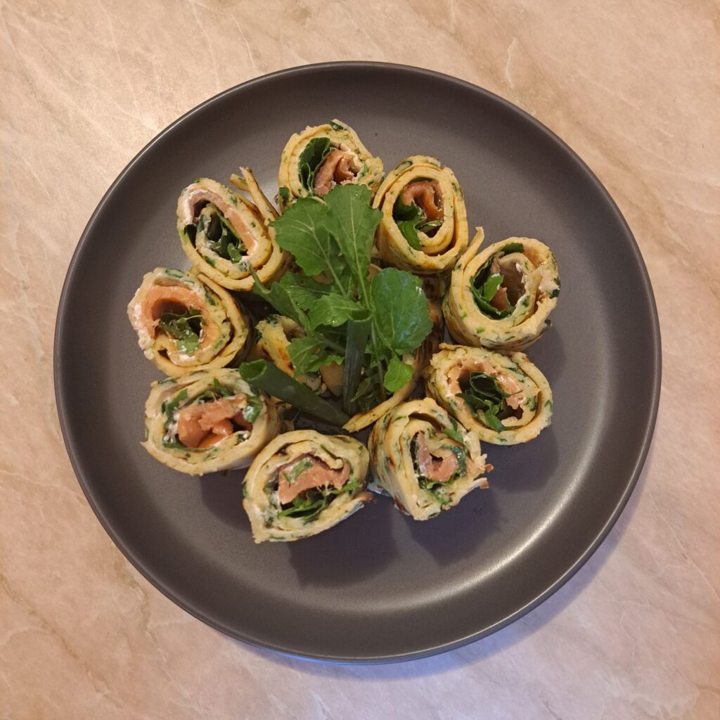 Smoked Salmon Savory Crepes sliced into rolls and arranged on a black plate, garnished with arugula leaves and whole spring onion greens.