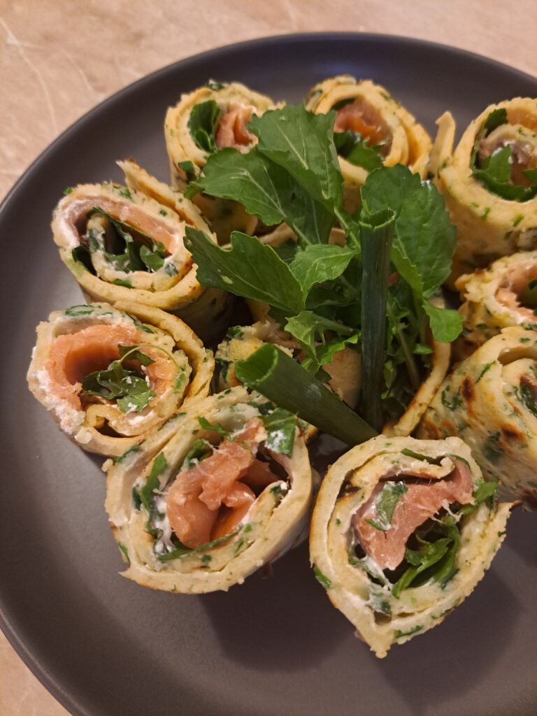 Rolled savory crepes filled with smoked salmon, sour cream, and arugula, sliced and arranged on a black plate, garnished with arugula leaves and green onion pieces.