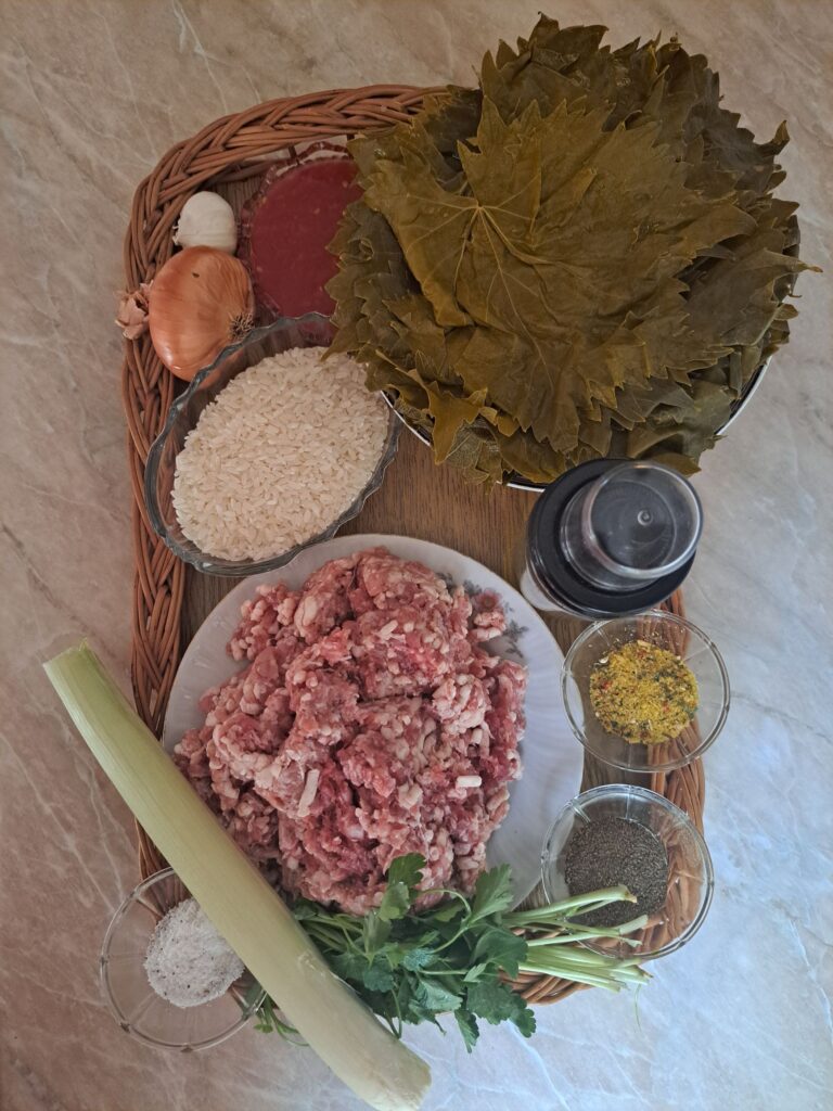 All ingredients for Beef Stuffed Grape Leaves arranged on a countertop, including grape leaves, ground beef, rice, tomato paste, onion, garlic, leek, parsley, spices, and cooking oil.