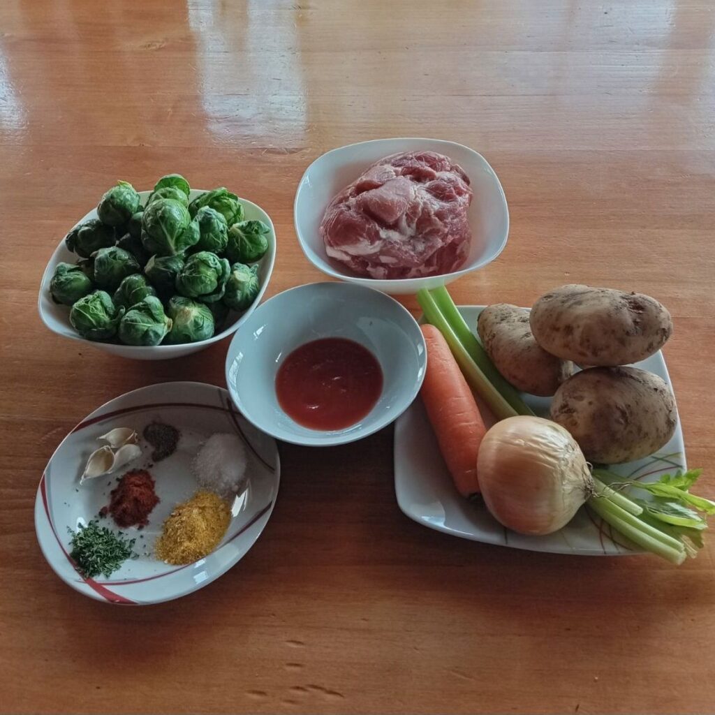 Chopped pork, potatoes, Brussels sprouts, onion, carrot, celery, garlic, and spices arranged neatly on a kitchen table.