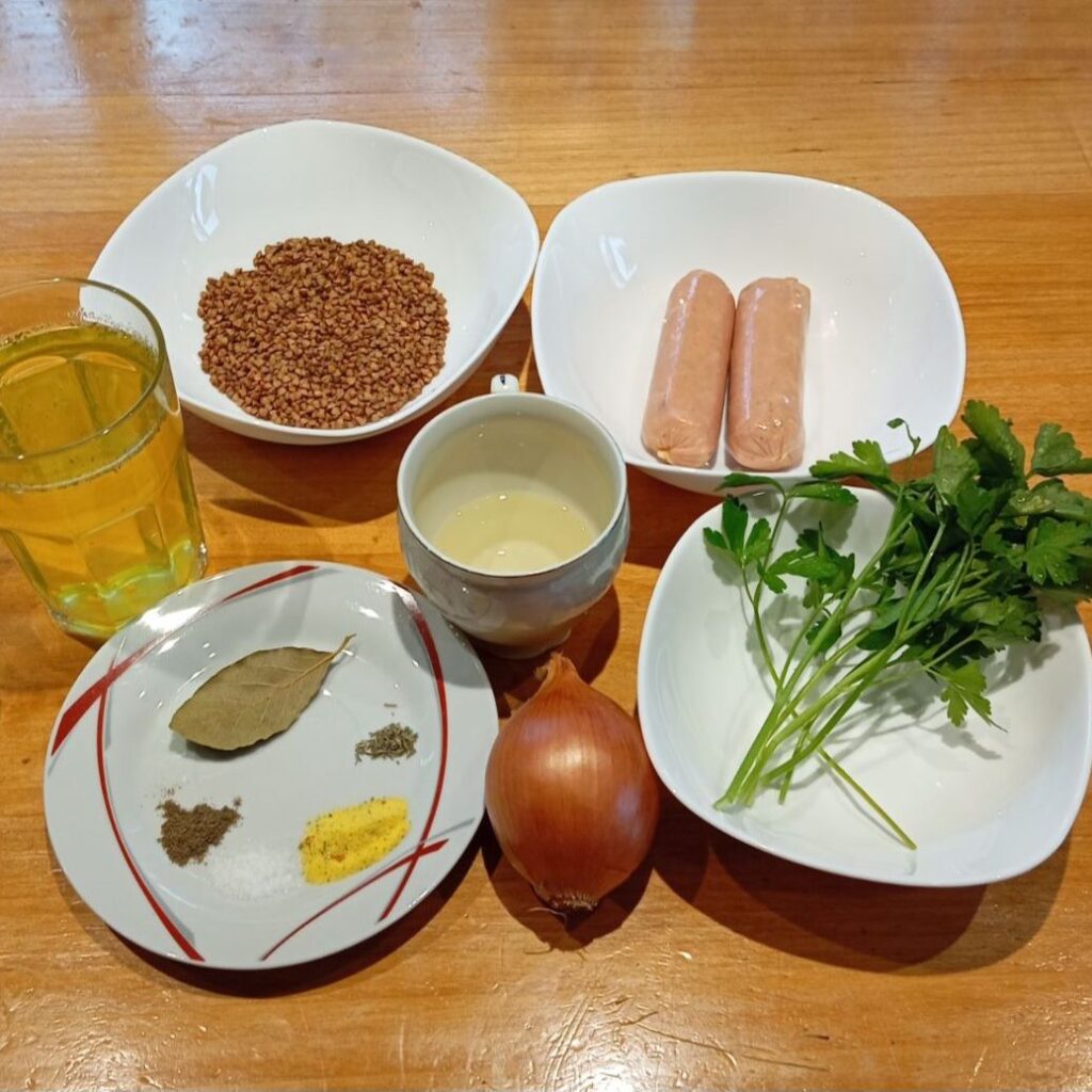 Fresh buckwheat, onion, parsley, pork sausages, seasonings, vegetable broth, and cooking oil arranged neatly on a wooden table.