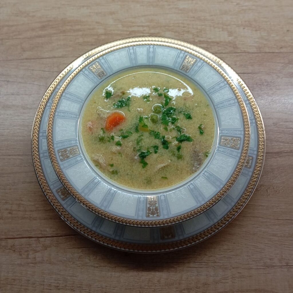 Deep wide plate of creamy white chicken soup served hot, with visible pieces of chicken, soft vegetables, smooth broth, and freshly chopped parsley on top.