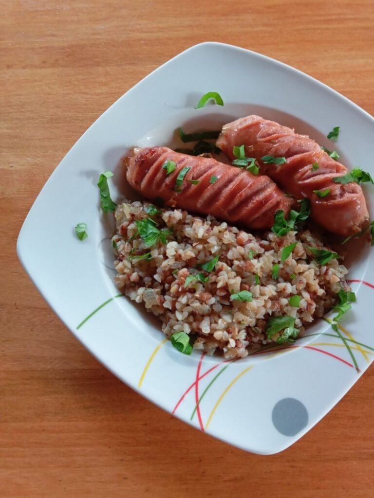 Close-up of a single portion of tender buckwheat with pork sausages served in a wide, deep plate, garnished with fresh parsley.