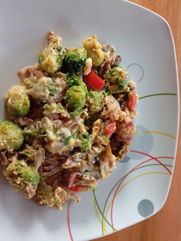 Close-up of Smoky Bacon Brussels Dish with crispy bacon, Brussels sprouts, mushrooms, red pepper, and melted cheese on a plate.