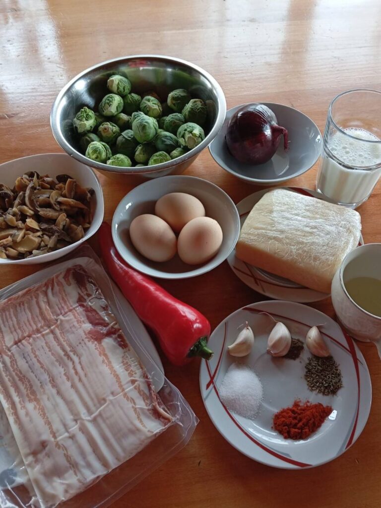 Fresh and prepared ingredients for Smoky Bacon Brussels Dish including bacon, Brussels sprouts, mushrooms, red pepper, onion, garlic, eggs, milk, and cheese.