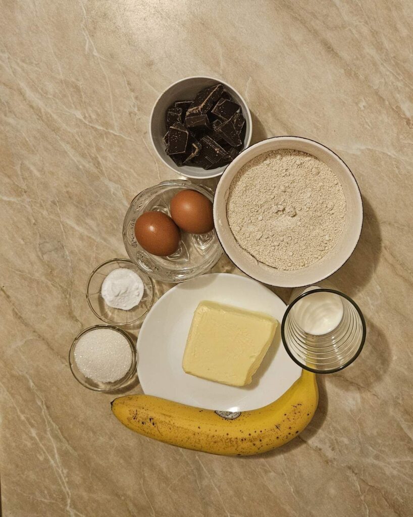 Measured ingredients for chocolate banana oat muffins arranged on a table including bananas, chocolate, flours, eggs, milk, butter, and sugar.
