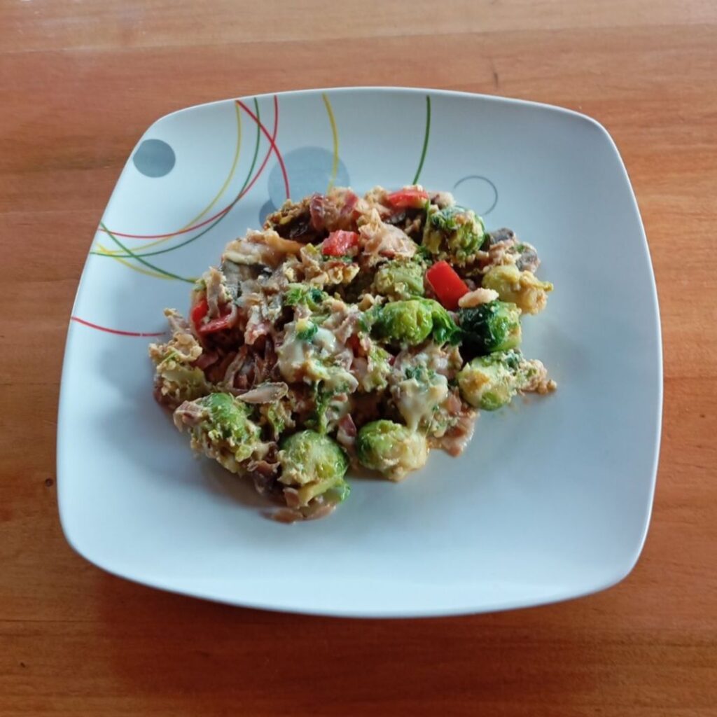 Smoky Bacon Brussels Dish served on a plate with bacon, Brussels sprouts, vegetables, and melted cheese.