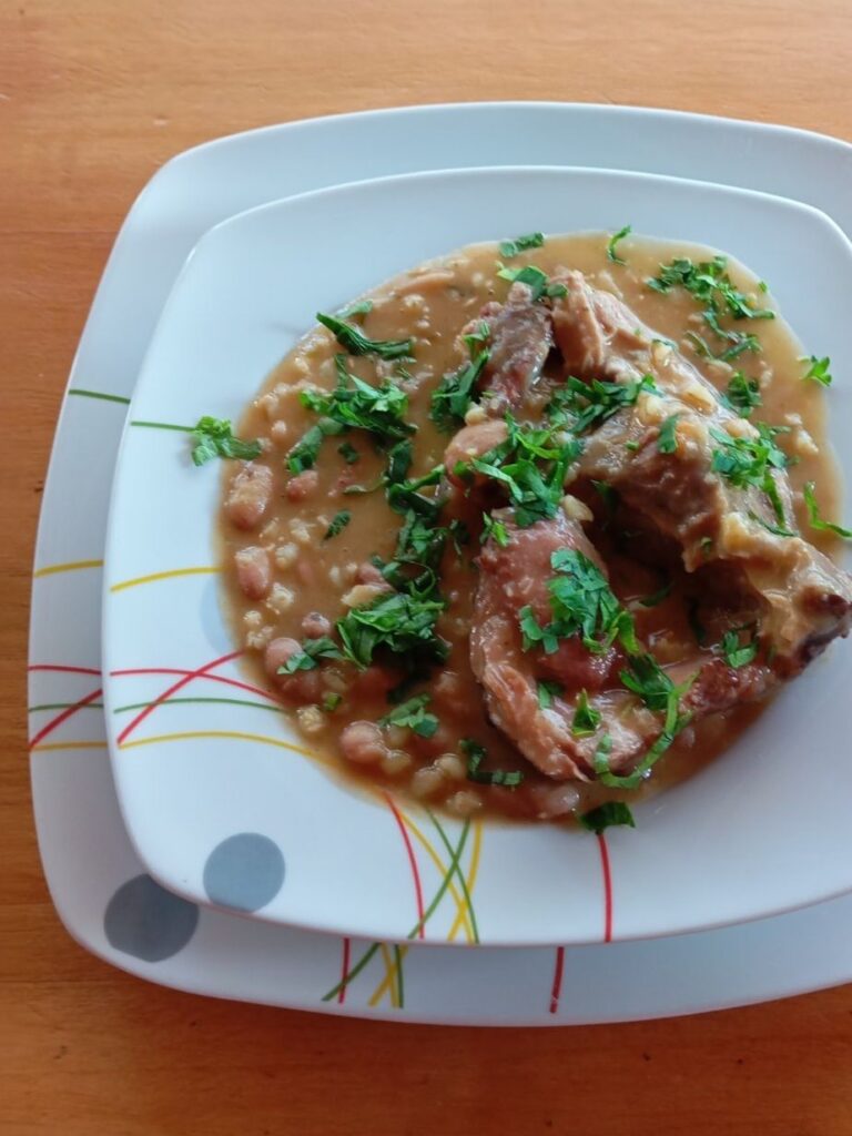 Close-up of bean soup with smoked mutton, barley, and vegetables in a deep plate, garnished with chopped parsley.