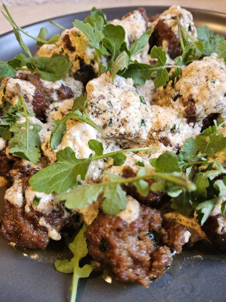 Small meatballs served in creamy white sauce on a flat black plate, garnished with fresh microgreens and arranged neatly for presentation.