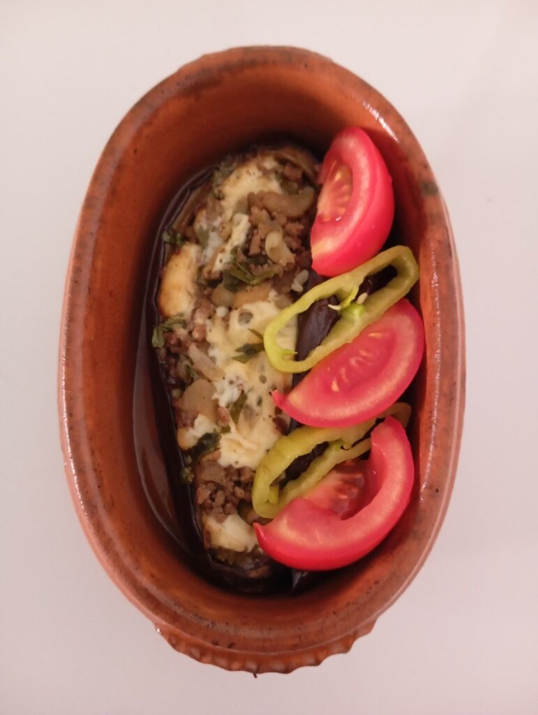 Close view of stuffed eggplant with minced meat, melted cheese, tomato pieces, and green pepper served in a small ceramic baking dish.