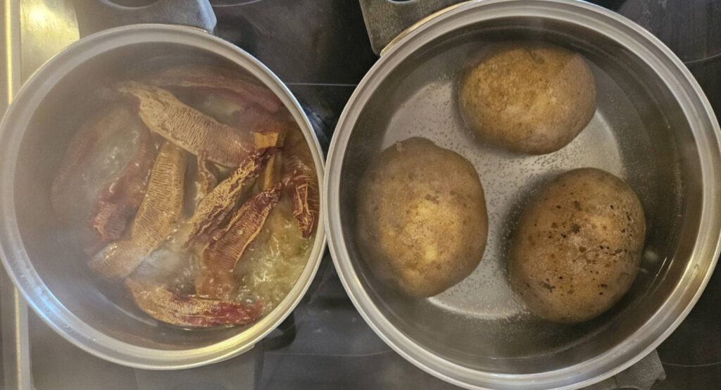 Two pots on a stovetop with whole potatoes boiling in one and dried peppers simmering in the other.