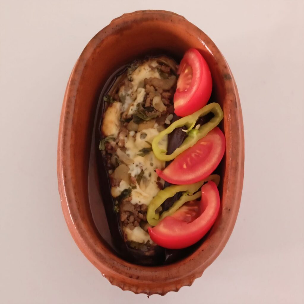 Served stuffed eggplant filled with minced meat and melted cheese in a ceramic baking dish with tomato pieces and green pepper slices on top.