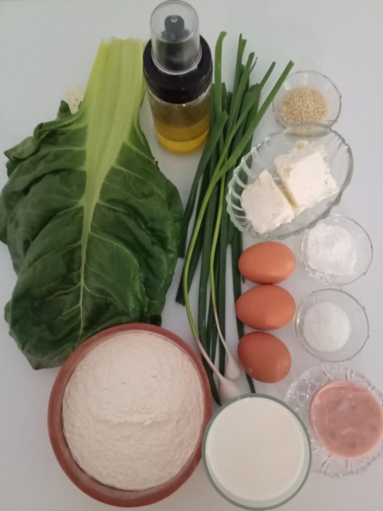 Flour, milk, cheese, ham, eggs, Swiss chard, spring onion, young garlic, sesame seeds, and baking ingredients arranged on a kitchen table.