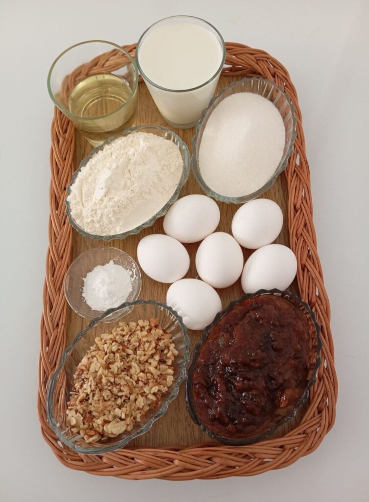 Measured ingredients for Fig Walnut Marshmallow Cake arranged on a kitchen table, including eggs, flour, sugar, milk, oil, fig marmalade, and chopped walnuts.