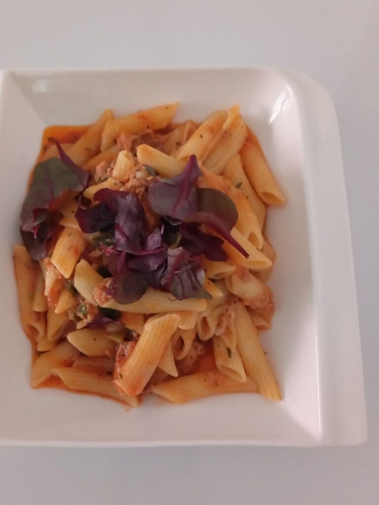 Close view of creamy tuna macaroni in a deep plate with tomato sauce, young onion, and fresh mixed microgreens on top.