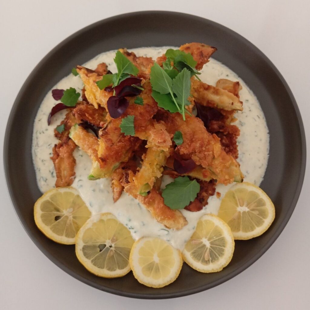 Served crunchy fried zucchini strips over creamy herb sauce with microgreens and lemon strips on a decorative plate.