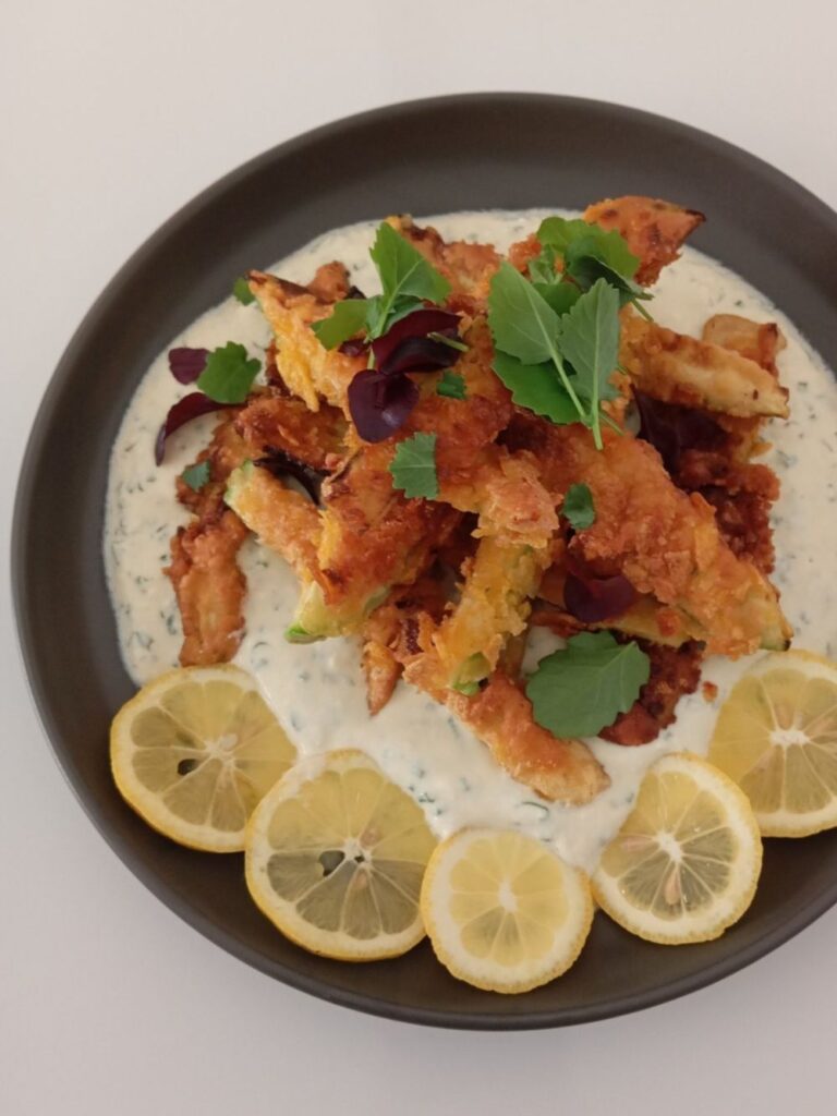 Close view of crunchy fried zucchini strips served over creamy herb sauce with microgreens and lemon slices on a plate.